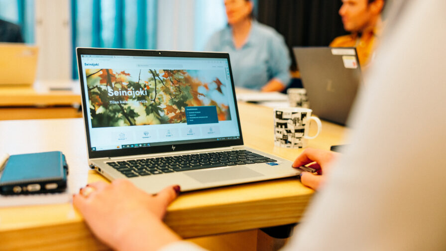 Kannettava tietokone auki henkilön edessä pöydällä. Taustalla kokouspöydän ympärillä myös muita ihmisiä.
