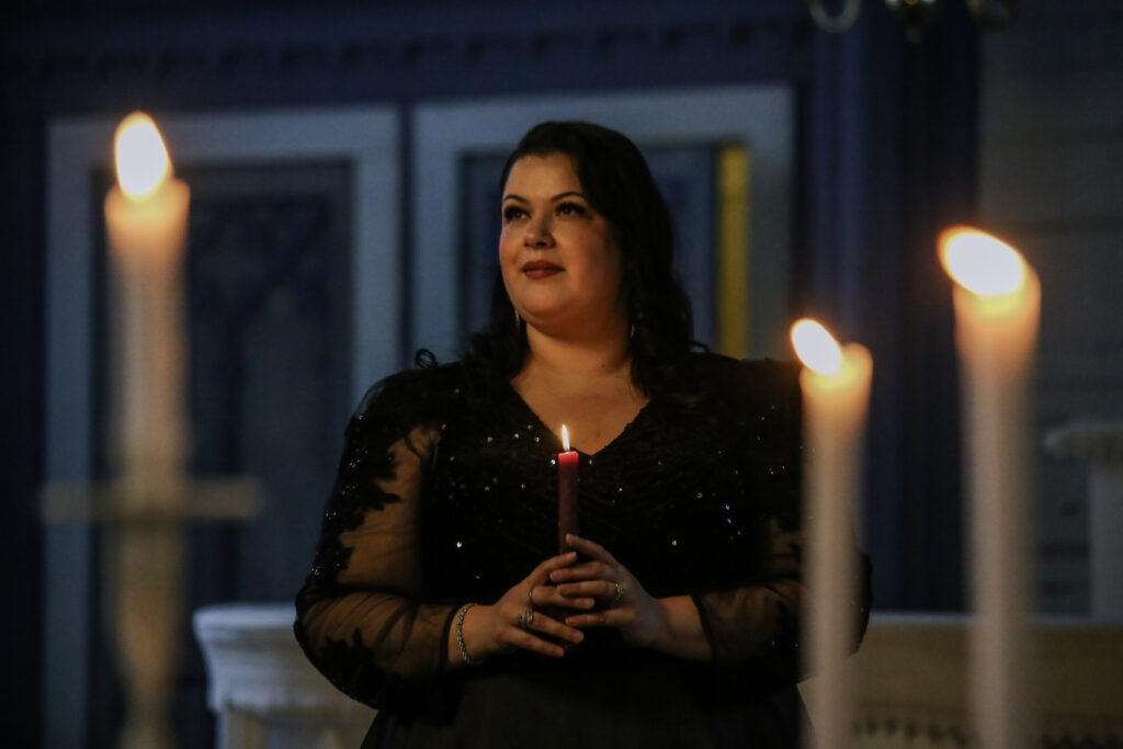 Nainen seisoo kirkossa ja pitää kynttilää käsissään. Etualalla kolme palavaa kynttilää.