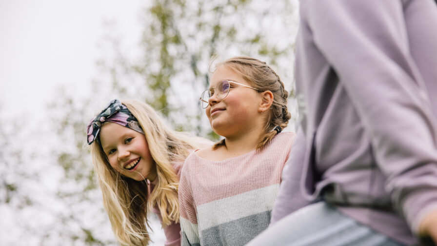 Lähikuvassa kolme lasta kiipeilytelineen päällä istumassa. Taustalla puiston puiden latvoja.