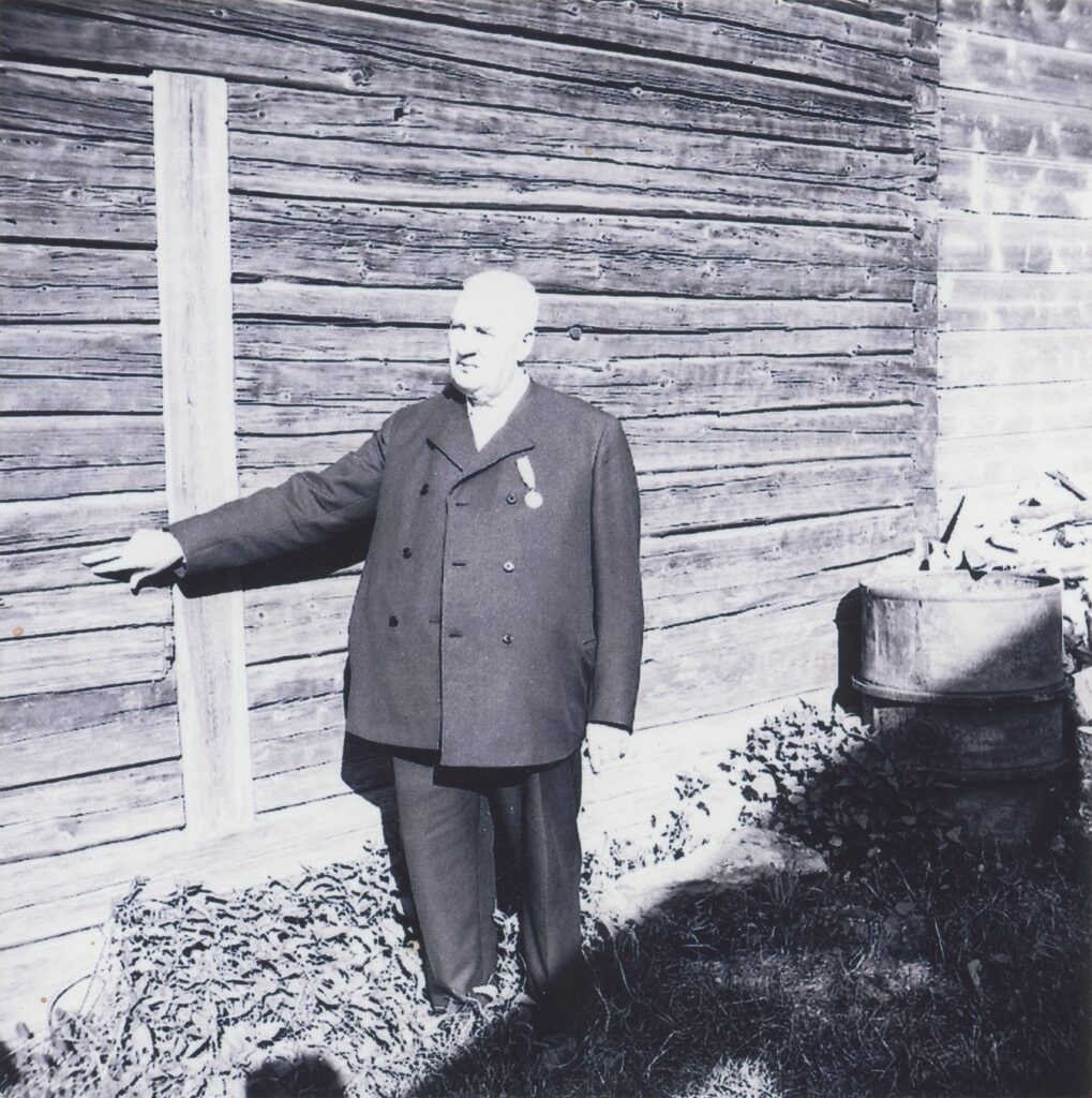 Mustavalkokuvassa mies seisoo osoittaen sormellaan hirsitalon seinää.