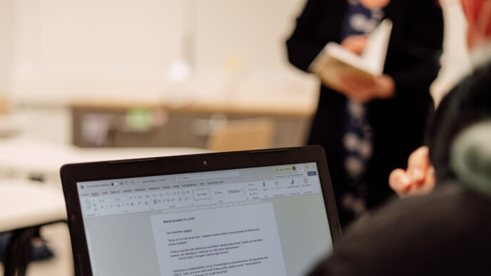 Kannettavan tietokoneen näyttö lähietäisyydeltä luokkahuoneessa, ja taustalla seisoo henkilö pitämässä kirjaa käsissään.