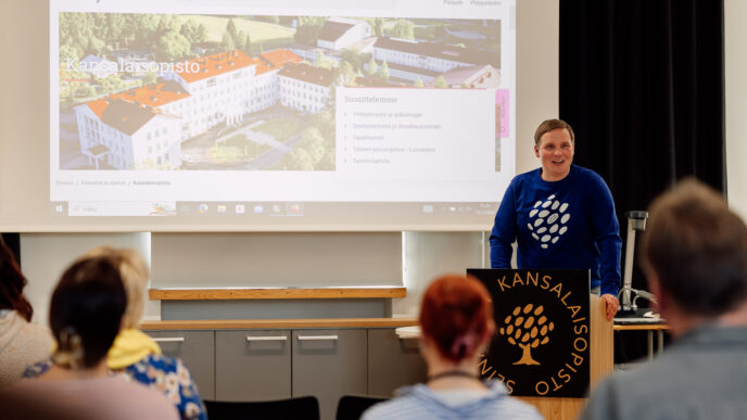 henkilö luentosalin edessä , taustalla valkokankaalle heijastettu kansalaisopiston nettisivu, kuvan etualalla näkyy henkilöitä katsomassa kohti huoneen etuosaa