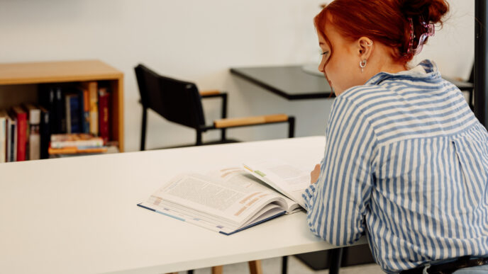 Henkilö istuu pöydän ääressä lukemassa avointa kirjaa rauhallisessa, kirjasto- tai opiskelutilaa muistuttavassa ympäristössä.