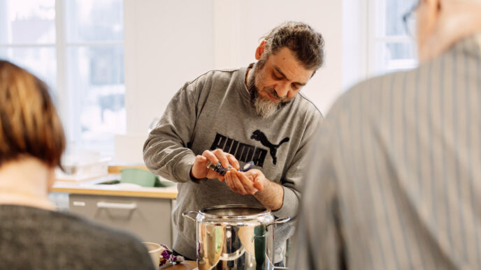 Henkilö valmistaa ruokaa keittiötilassa, sekoittaen aineksia teräskattilaan, kun kaksi muuta henkilöä seuraa työskentelyä läheltä.