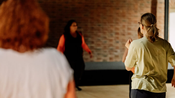 Ihmisryhmä harjoittelee liikkumista tai tanssia avarassa sisätilassa, jossa on tiiliseinä ja puulattia