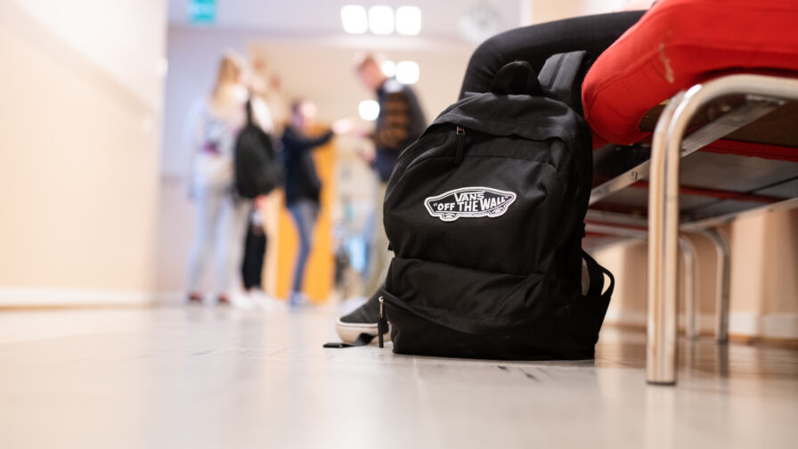 Reppu nojaa penkkiä koulun käytävällä. taustalla seisoskelee opiskelijoita.