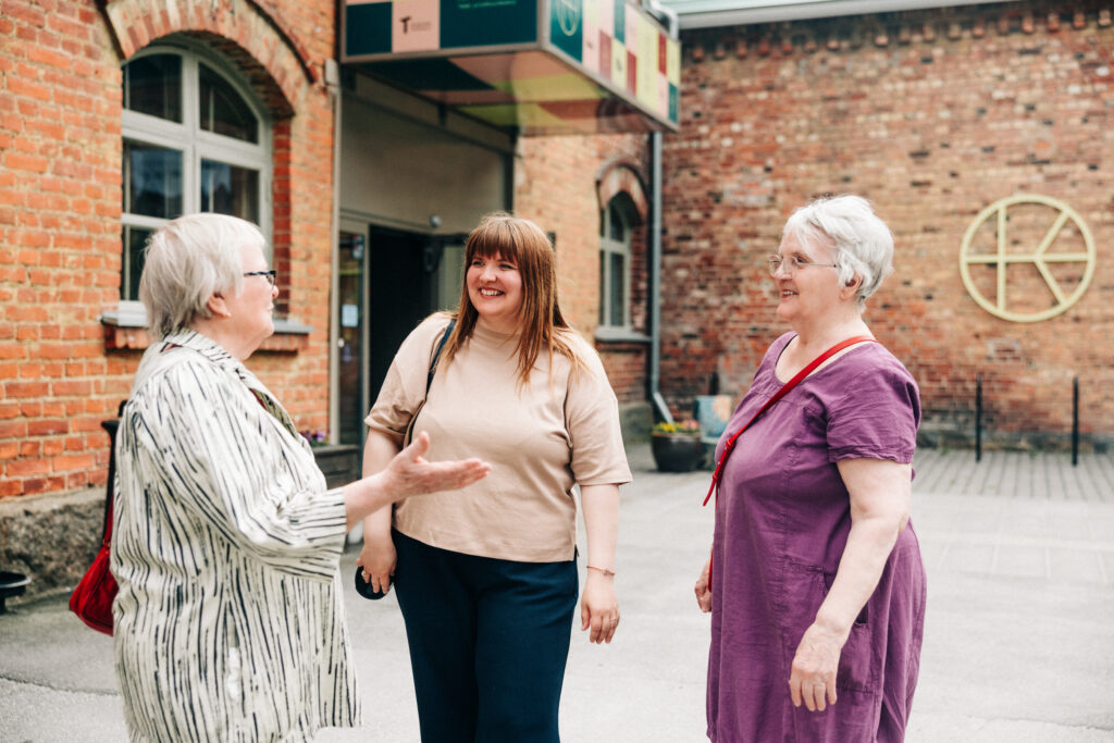 Kolme henkilöä seisoo ulkona tiilirakennuksen edessä katsoen toisiaan hymyillen