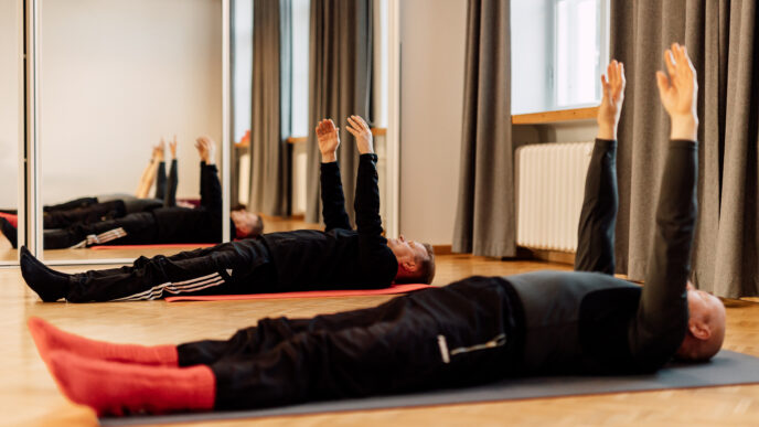 Ihmiset makoilevat joogamatolla selällään jumppasalissa, kädet suorina kohti kattoa peilien edessä.