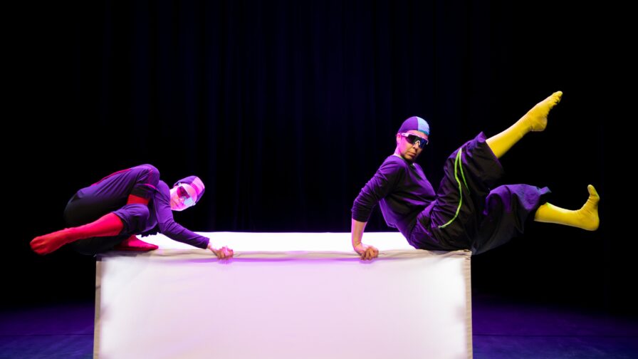 kaksi henkilöä tasapainoilee isoa laatikkoa muistuttavan esineen päällä värikkää esiintymisasut yllään.