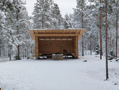 Kuvassa on Karhuvuoren laavu ja nuotiopaikka talvisessa metsässä. Kuvassa on lumisia puita. 