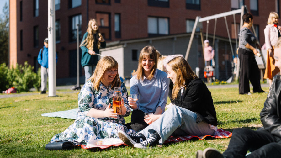 Kolme henkilöä istuu piknikillä nurmikolla kerrostalon pihalla. Taustalla kaikenikäisiä ihmisiä keinumassa, leikkimässä ja juttelemassa.