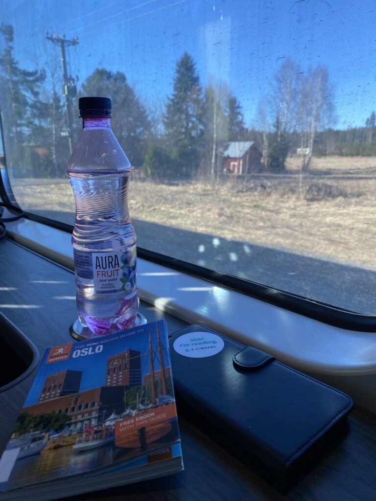 Näkymä junan ikkunasta, ikkunan edessä pöydällä Oslon matkaopas, vesipullo ja puhelin.