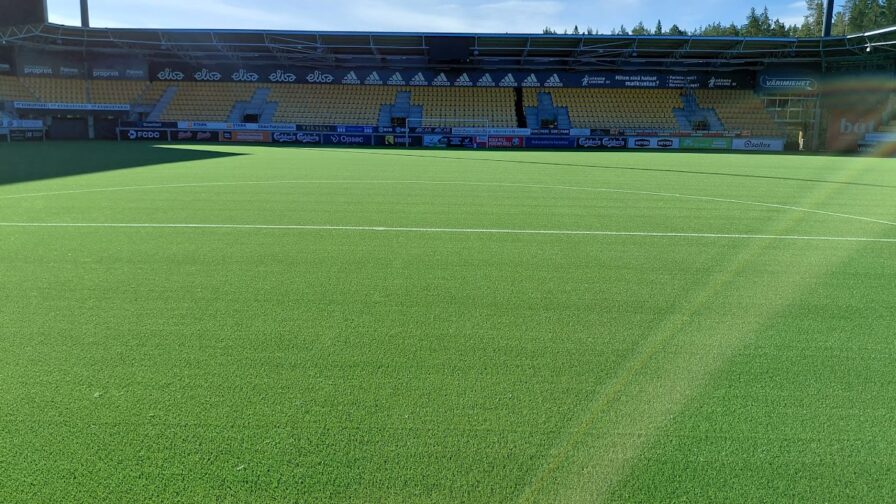 Vihreä tekonurmikenttä. Taustalla jalkapallostadionin katsomo.