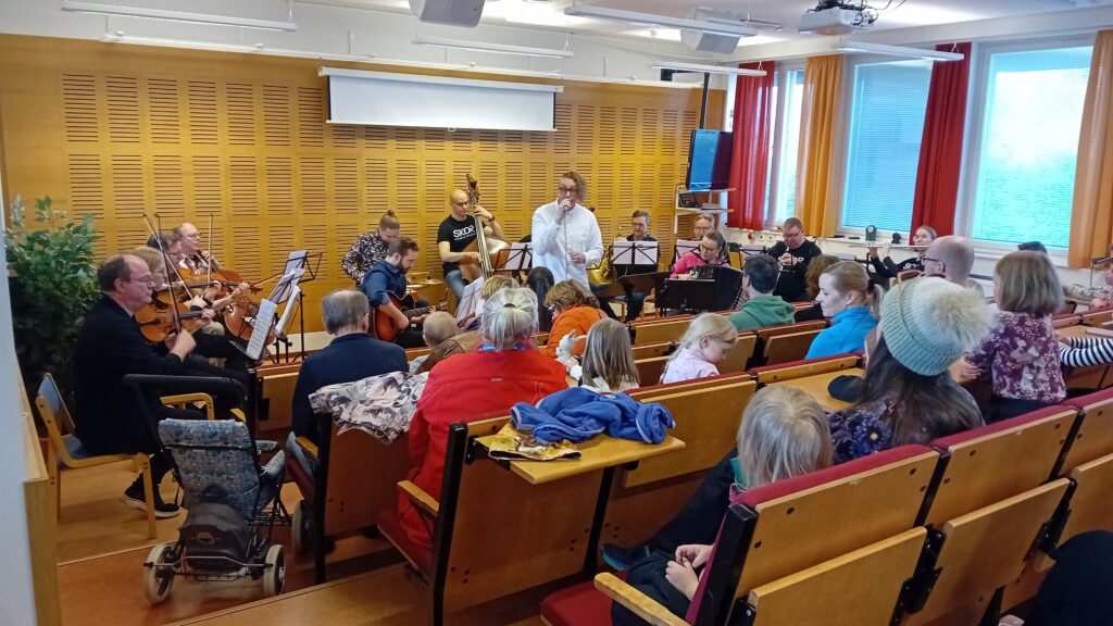 Auditorio täynnä lapsiperheitä, edessä muusikoita soittamassa ja laulamassa.
