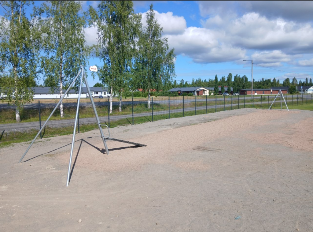 Kuvassa on vaijeriliuku, joka on asennettu hiekkakentälle. Taustalla näkyy aitaa, tietä, puita ja asuinrakennuksia.