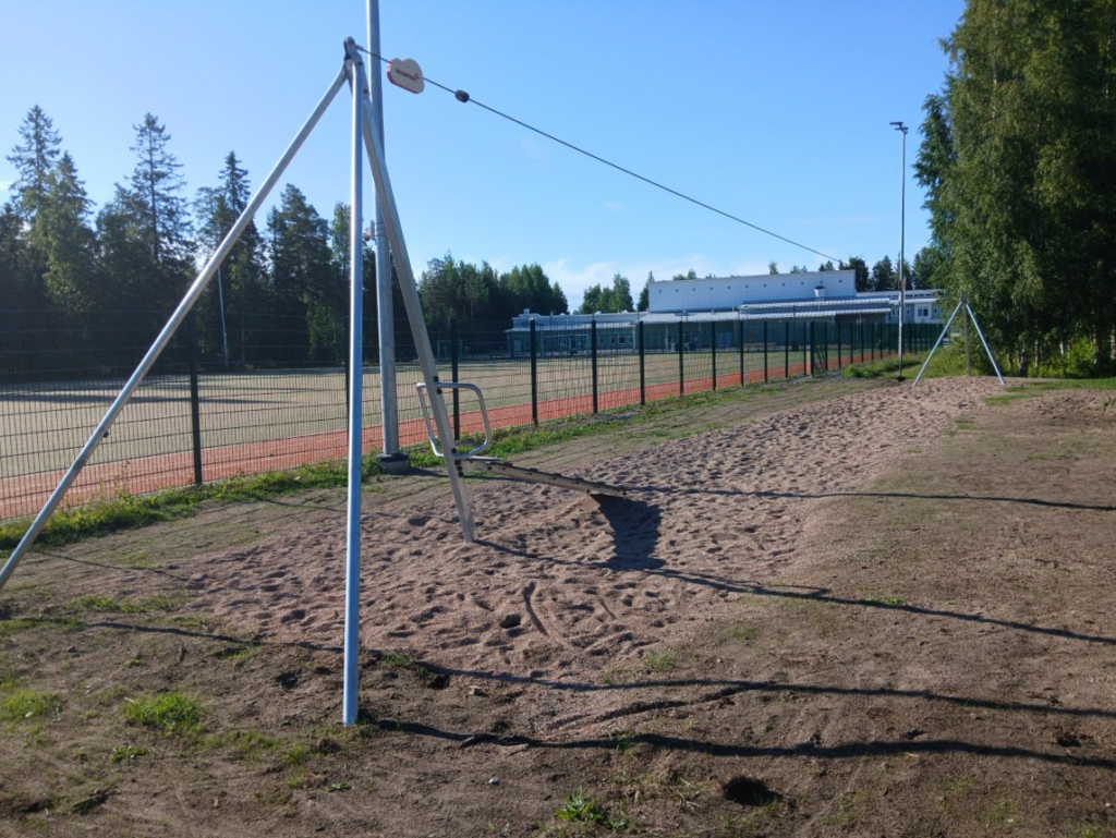Kuvassa on vaijeriliuku joka on asennettu urheilukentän vierelle. Taustalla näkyy koulurakennus.