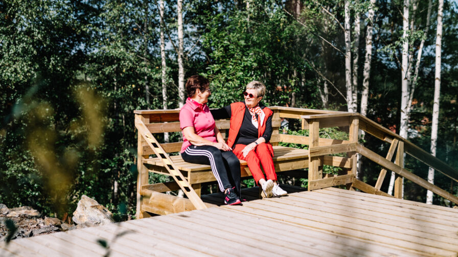 Kaksi ikääntynyttä henkilöä istuu elämysportaiden penkillä ja juttelee keskenään. Taustalla lehtipuita.