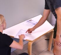 kaksi henkilöä pöydän edessä ja osoittavat pöydällä olevia papereita.